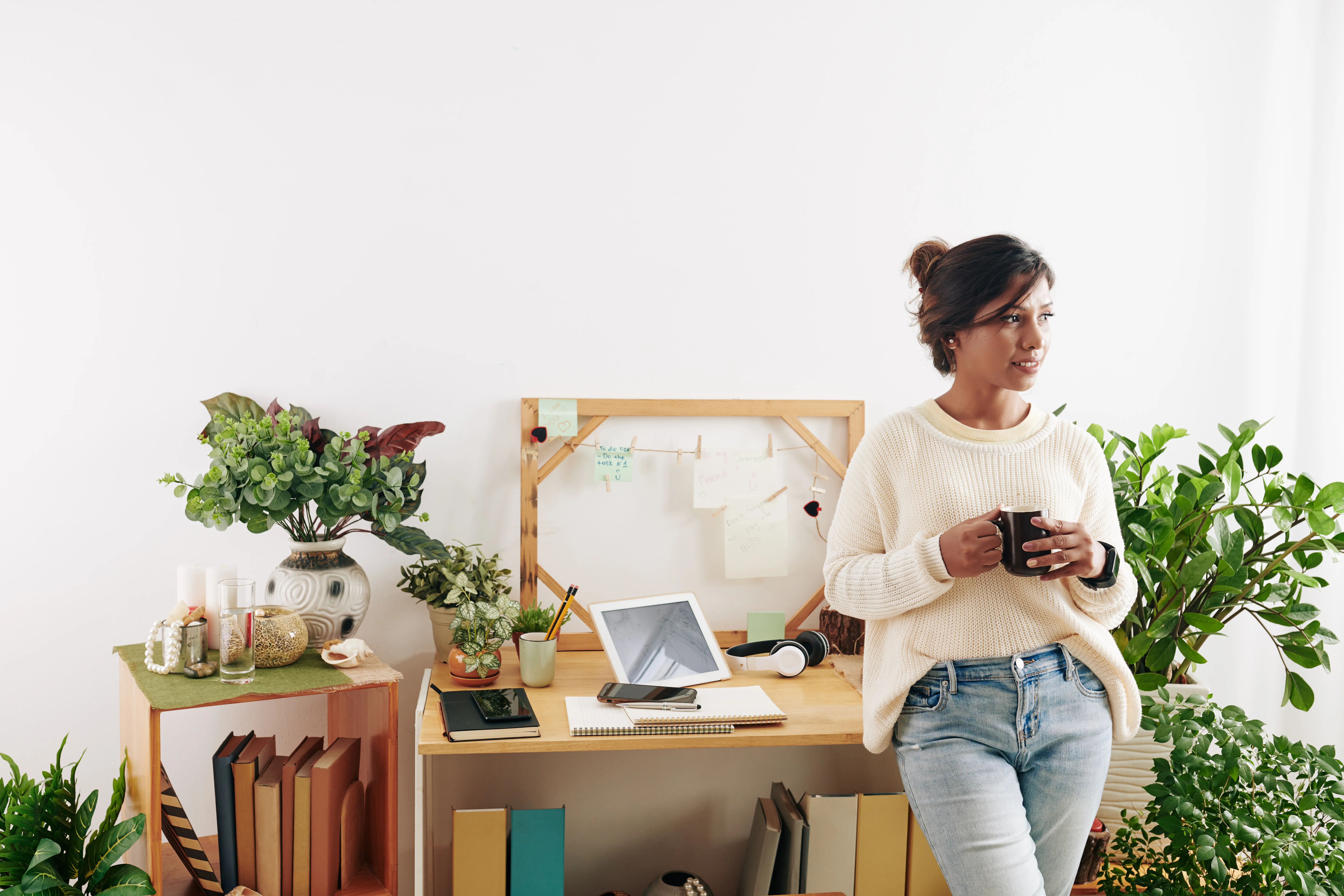 Porträt einer jungen, eleganten Grafikdesignerin, die am Schreibtisch steht, eine Tasse Kaffee in den Händen hält und wegschaut.